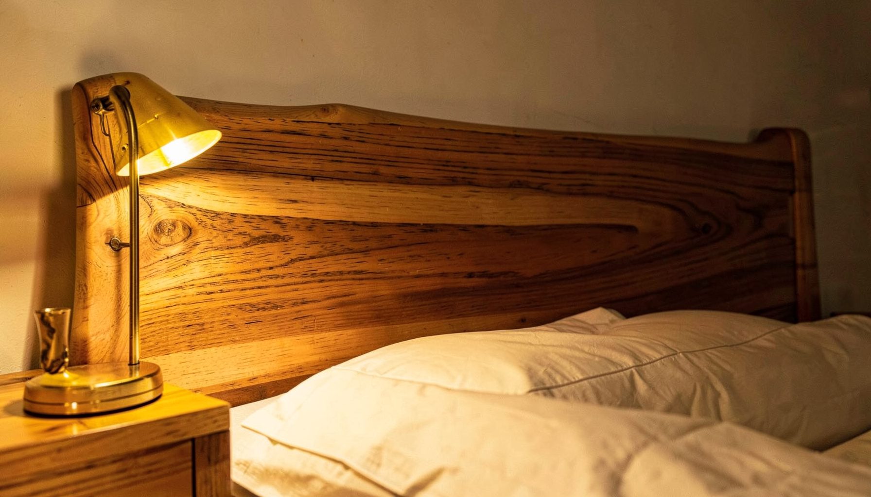 Dettaglio di una testiera di letto in legno di noce con venature naturali a vista, illuminata lateralmente da una lampada da comodino che crea un'atmosfera calda e raccolta.