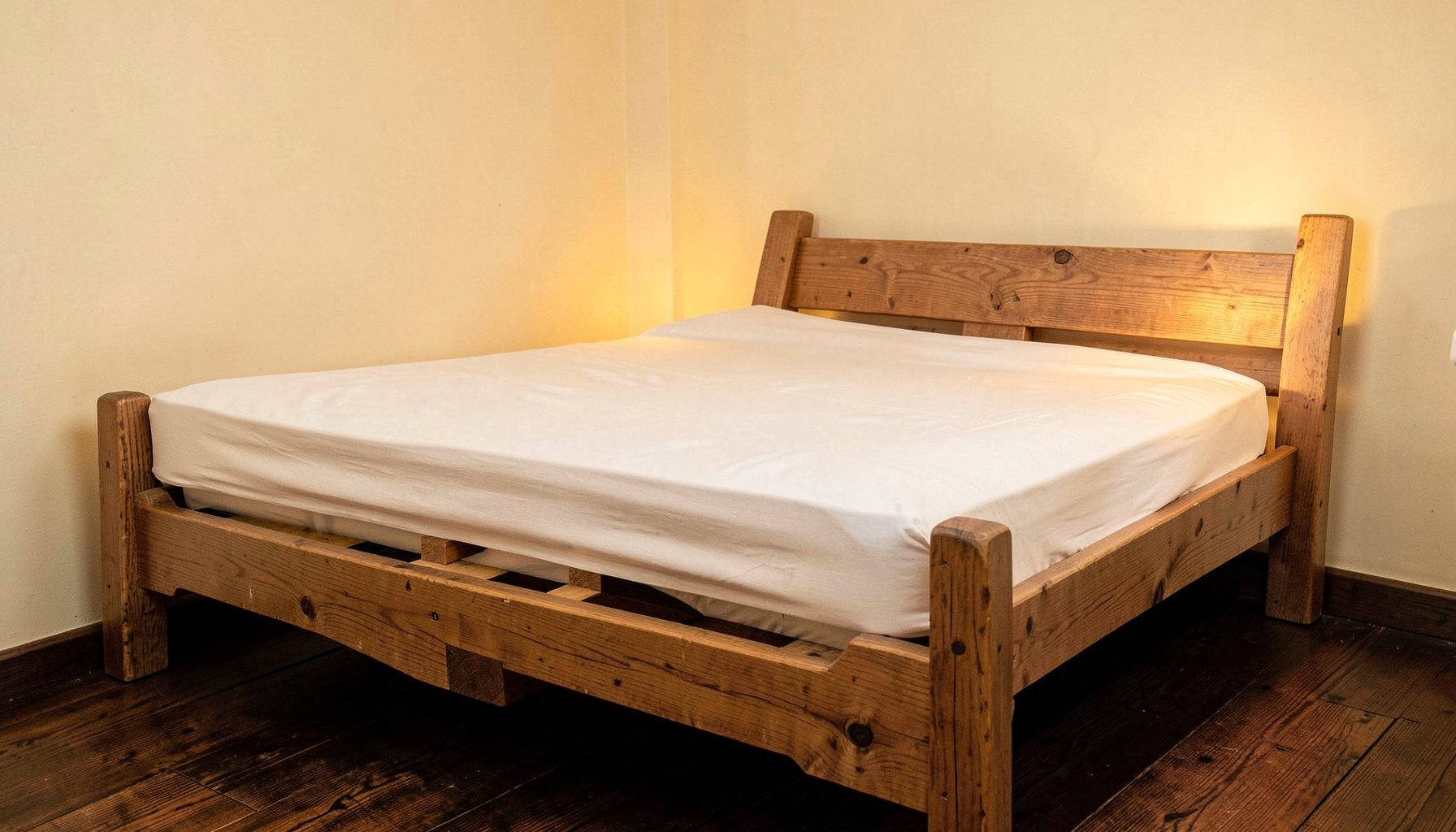 Un letto matrimoniale in legno naturale con materasso bianco e rete a doghe visibile, illuminato da luce calda, in una camera dalle pareti color crema e pavimento in legno scuro